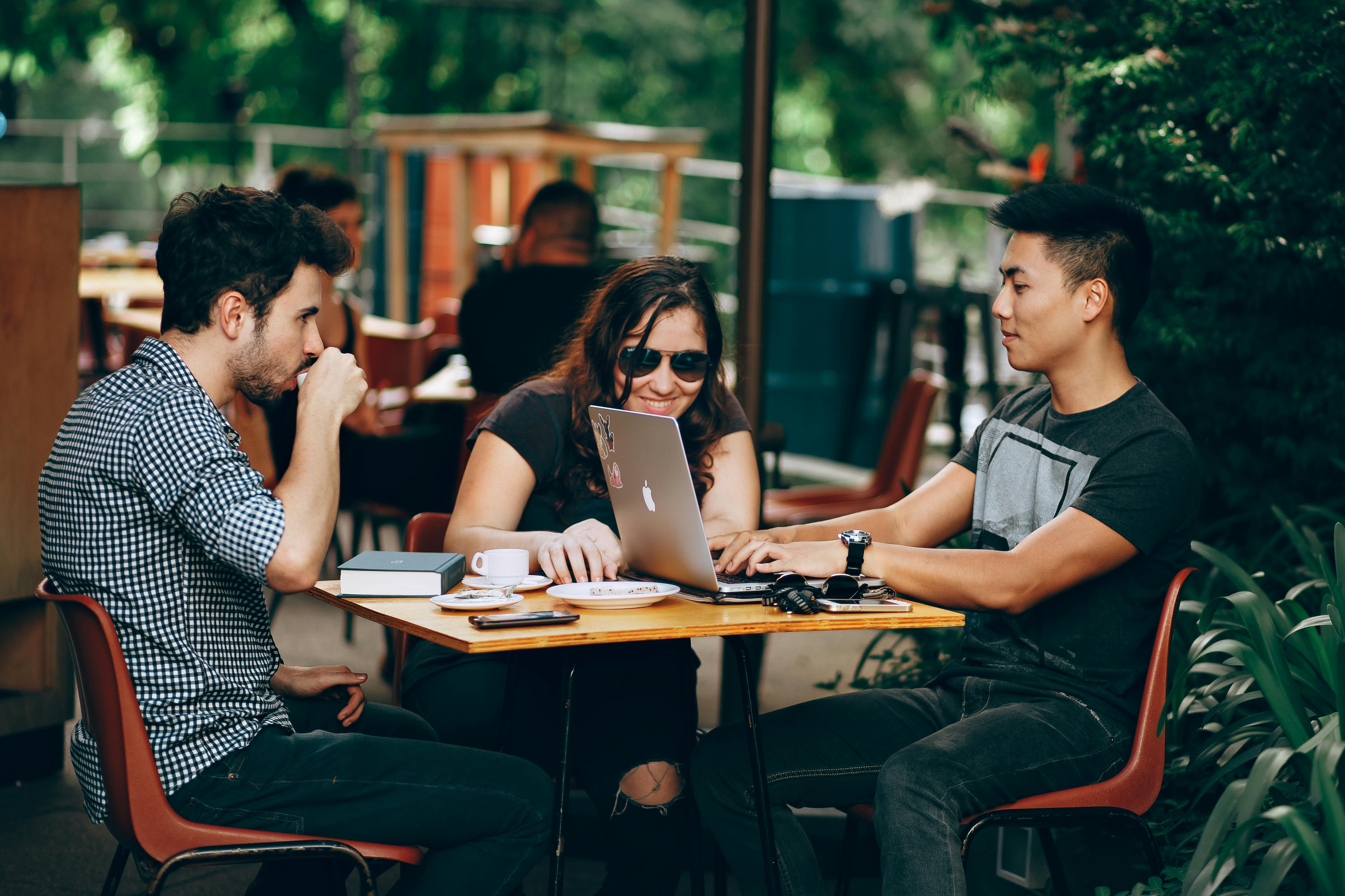 a group of three young people sitting together at a table outdoors. One person is on a laptop, the second is looking at the laptop screen, and the third is drinking from a cup