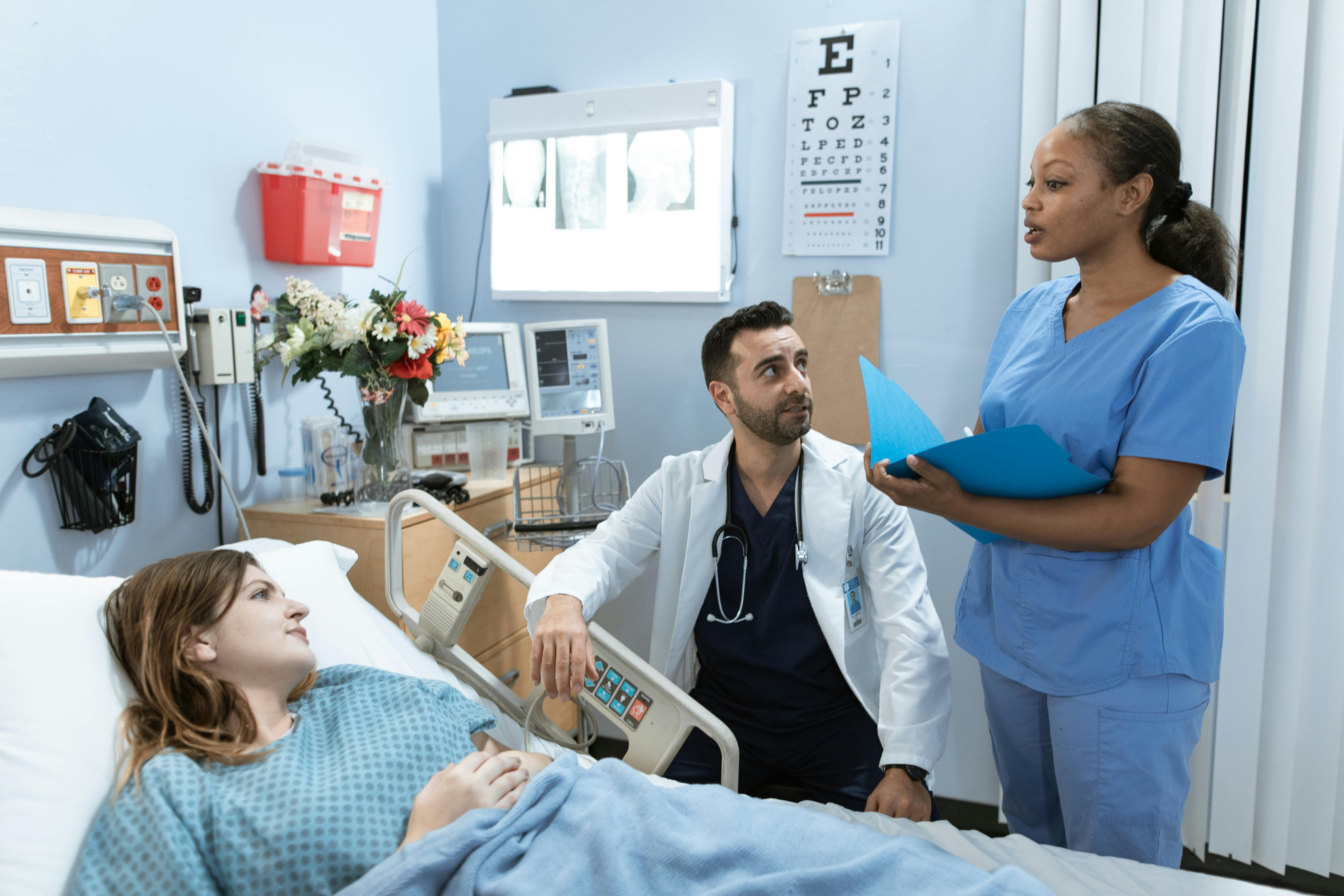 a nurse speaking with a patient in a hospital bed while a doctor sits on a stool and observes